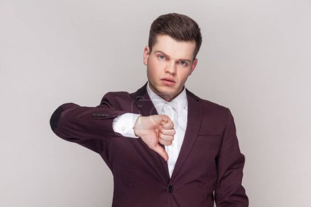 Photo for Portrait of handsome man shows disapproval sign, keeps thumb down, expresses dislike, frowns face in discontent, wearing violet suit and white shirt. Indoor studio shot isolated on grey background. - Royalty Free Image