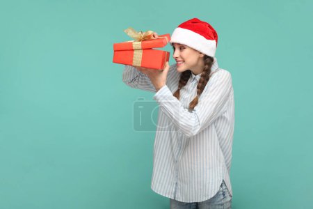 Foto de Retrato de chica optimista feliz con trenzas con camisa a rayas y sombrero de Santa Claus, sosteniendo la caja de regalo, mirando dentro con sonrisa. Estudio interior plano aislado sobre fondo verde. - Imagen libre de derechos
