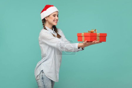 Photo for Side view portrait of smiling delighted teenager girl wearing striped shirt and Santa Claus hat, congratulating with new year, giving present. Indoor studio shot isolated on green background. - Royalty Free Image