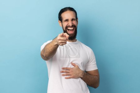 Photo for Well you're a joker. Portrait of positive handsome man with beard wearing white T-shirt pointing index finger at camera and laughing positively. Indoor studio shot isolated on blue background. - Royalty Free Image