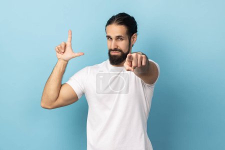 Téléchargez les photos : Portrait d'un homme frustré barbu portant un T-shirt blanc montrant un geste plus lâche et pointant vers la caméra, regardant avec un visage grincheux. Studio intérieur tourné isolé sur fond bleu. - en image libre de droit