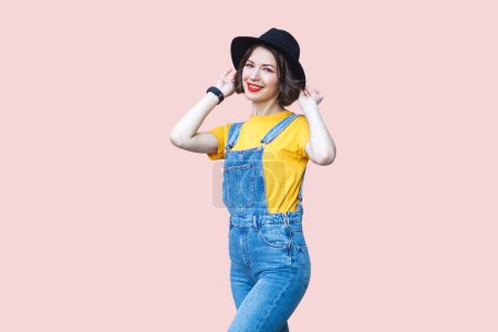 Téléchargez les photos : Portrait d'une jolie femme hipster satisfaite en salopette en denim bleu, T-shirt jaune, garde la main sur son chapeau noir, souriant à la caméra. Studio intérieur isolé sur fond rose clair. - en image libre de droit