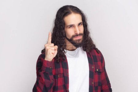 Photo for Attention please. Portrait of serious bearded man with long curly hair in checkered red shirt raises finger up, warning, has confident look. Indoor studio shot isolated on gray background. - Royalty Free Image