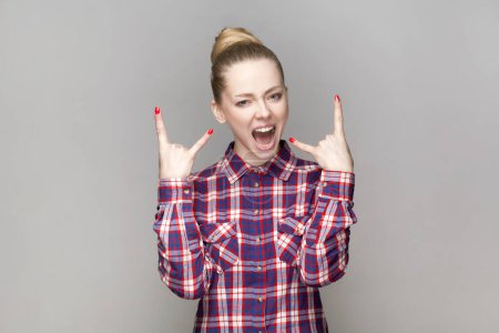 Photo for Portrait of crazy excited beautiful woman with bun hairstyle standing showing rock and roll gesture, screaming, wearing checkered shirt. Indoor studio shot isolated on gray background. - Royalty Free Image