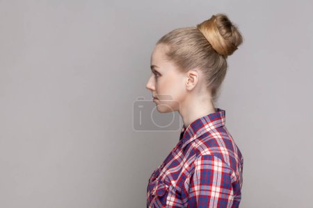 Photo for Side view portrait of serious self-confident woman with bun hairstyle standing looking ahead with concentrated facial expression wearing checkered shirt. Indoor studio shot isolated on gray background - Royalty Free Image