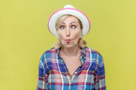Photo for Portrait of mature woman wearing checkered shirt and hat, puckers lips makes grimace, imitates fish being childish does facial expressions. Indoor studio shot isolated on yellow background. - Royalty Free Image