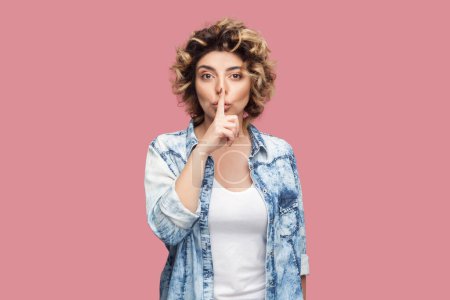 Photo for Portrait of serious beautiful woman with curly hairstyle wearing blue shirt standing with finger near lips, showing shh gesture, keep silence. Indoor studio shot isolated on pink background. - Royalty Free Image