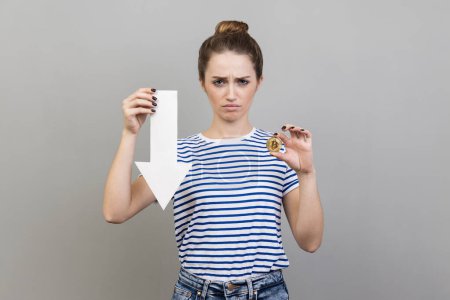 Photo for Portrait of sad upset woman wearing striped T-shirt showing of bitcoin and white arrow pointing down, expressing negative emotions. Indoor studio shot isolated on gray background. - Royalty Free Image