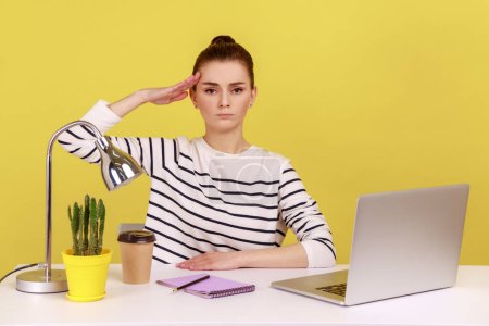 Photo for Yes sir. Obedient responsible woman office manager sitting at workplace with laptop and saluting, listening to boss order, corporate discipline. Indoor studio studio shot isolated on yellow background - Royalty Free Image