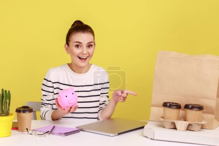 Photo for Friendly woman holding piggybank and pointing at coffee with pizza box, looking at camera with positive expression, sitting on workplace. Indoor studio studio shot isolated on yellow background. - Royalty Free Image