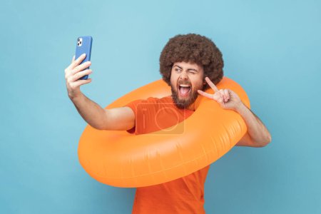 Photo for Man with Afro hairstyle wearing orange T-shirt holding orange rubber ring and cell phone, having video call or broadcasting livestrem showing v sign. Indoor studio shot isolated on blue background. - Royalty Free Image