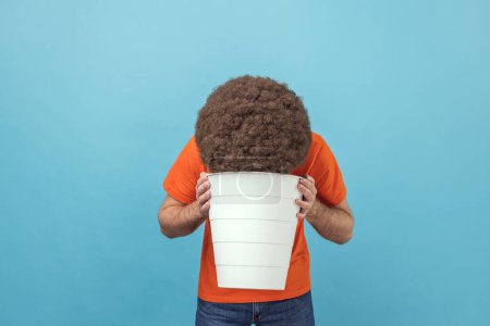 Photo for Portrait of sick unhealthy man with Afro hairstyle wearing orange T-shirt standing with white bucket, vomiting, suffering bad feelings, discomfort. Indoor studio shot isolated on blue background. - Royalty Free Image