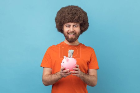 Photo for Portrait of smiling man with Afro hairstyle wearing orange T-shirt holding piggy bank with dollar banknote in hands, being happy of savings. Indoor studio shot isolated on blue background. - Royalty Free Image