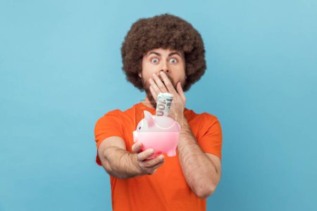 Photo for Portrait of shocked man with Afro hairstyle wearing orange T-shirt holding piggy bank with dollar banknote in hands, being surprised of savings. Indoor studio shot isolated on blue background. - Royalty Free Image