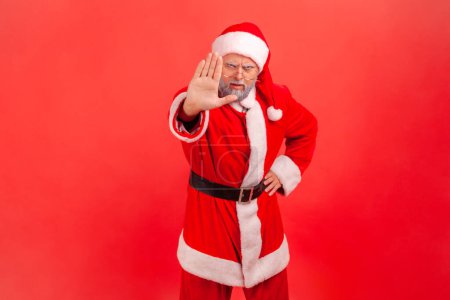 Photo for Portrait of serious elderly man with gray beard wearing santa claus costume showing stop ban gesture with palm, sign of prohibition, with hand. Indoor studio shot isolated on red background. - Royalty Free Image