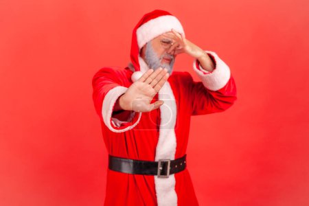 Téléchargez les photos : Père Noël en costume rouge pincer le nez avec les doigts pour éviter les mauvaises odeurs et montrer un geste d'arrêt, démontrant la répulsion à puer, odeur terrible. Studio intérieur tourné isolé sur fond rouge. - en image libre de droit