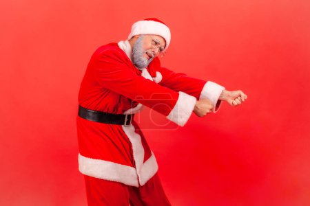 Photo for Elderly man with gray beard in santa claus costume pretending to pull invisible rope, concept of hard working, striving and efforts to achievements. Indoor studio shot isolated on red background. - Royalty Free Image
