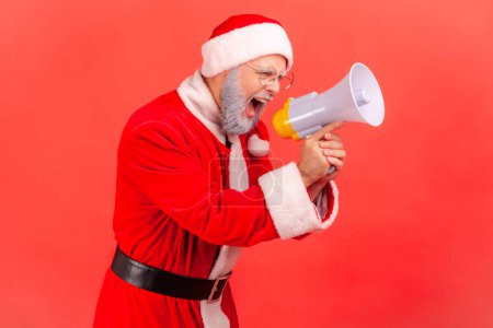 Photo for Side view of angry elderly man with gray beard wearing santa claus costume standing with megaphone in hands, shouting announcement, serious expression. Indoor studio shot isolated on red background. - Royalty Free Image
