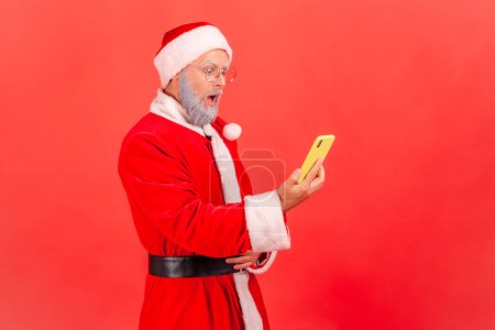 Téléchargez les photos : Vue latérale de l'homme âgé avec barbe grise portant le costume de Père Noël debout avec la bouche ouverte et tenant le téléphone intelligent, voit le contenu étonné. Studio intérieur tourné isolé sur fond rouge. - en image libre de droit