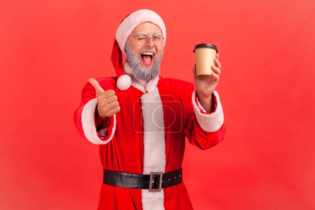 Photo for Portrait of happy elderly man with gray beard wearing santa claus costume standing with disposable paper cup with coffee, showing thumb up. Indoor studio shot isolated on red background. - Royalty Free Image