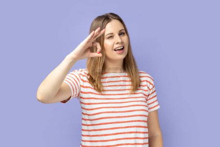 Photo for Portrait of cheerful young adult blond woman wearing striped T-shirt showing okay sign, assures you everything is fine, looks gladly. Indoor studio shot isolated on purple background. - Royalty Free Image