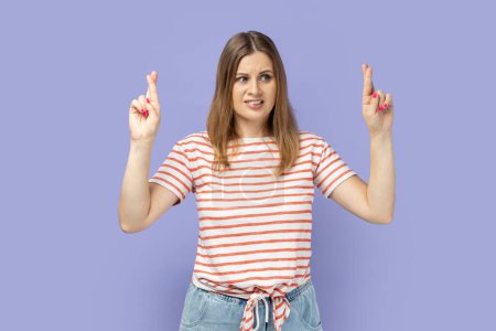 Photo for Portrait of hopeful joyous attractive blond woman wearing striped T-shirt raising fingers crossed while making wish, confident to win. Indoor studio shot isolated on purple background. - Royalty Free Image
