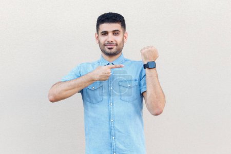 Photo for Portrait of handsome optimistic man wearing denim shirt pointing at his smart watch, looking at camera, warning about deadline. Indoor studio shot isolated on gray background. - Royalty Free Image