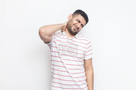 Photo for Portrait of handsome sick tired bearded man wearing striped t-shirt standing massaging his painful neck, frowning face from terrible pain. Indoor studio shot isolated on gray background. - Royalty Free Image