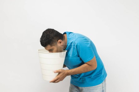 Photo for Side view portrait of sick unshaven man wearing blue T- shirt standing suffering stomachache, feels nausea and vomits, holding bin in hands. Indoor studio shot isolated on gray background. - Royalty Free Image