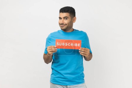 Photo for Portrait of smiling optimistic unshaven man in blue T- shirt standing holding card with subscribe inscription, looking at camera with happy expression. Indoor studio shot isolated on gray background. - Royalty Free Image