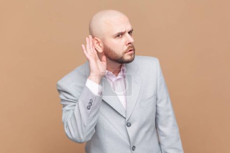 Photo for Portrait of bald man overhearing, listening intently to secret information, private talk, holding hand near ear to hear better, wearing gray jacket. Indoor studio shot isolated on brown background. - Royalty Free Image