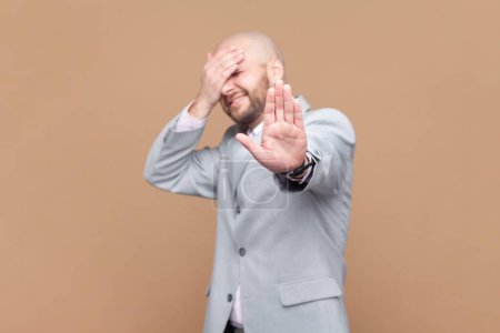 Photo for Portrait of scared frighten bald bearded man covering his face and showing stop hand gesture, sees something shameful, wearing gray jacket. Indoor studio shot isolated on brown background. - Royalty Free Image
