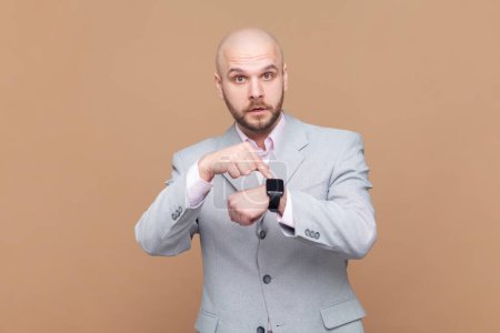 Photo for You are late. Portrait of serious bossy bald bearded man standing, looking at camera and pointing at his smart watch, wearing gray jacket. Indoor studio shot isolated on brown background. - Royalty Free Image