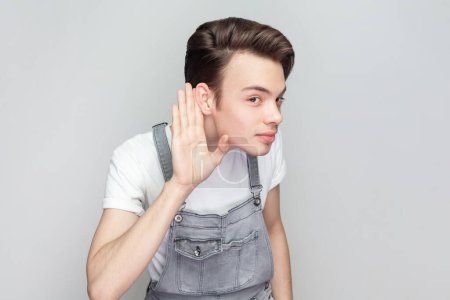 Photo for Intrigued curious young brunette man standing keeps hand near ear tries to overhear interesting conversation, looks attentively, wearing denim overalls. Indoor studio shot isolated on gray background. - Royalty Free Image