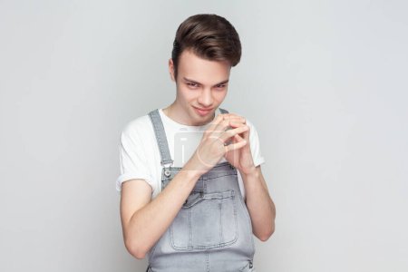 Photo for My plan is perfect. Young brunette man schemes something, steepls fingers and looks with cunning expression aside, smiles sly, wearing denim overalls. Indoor studio shot isolated on gray background. - Royalty Free Image