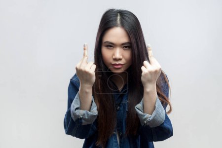 Foto de Portrait of brunette woman in blue denim jacket standing shows fuck you sign, looks with serious face, being vulgar and has quarrel with someone. Indoor studio shot isolated on gray background. - Imagen libre de derechos