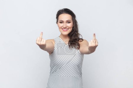 Téléchargez les photos : Portrait of brunette woman showing fuck you sign, looking with smile, being vulgar and has quarrel with someone, wearing striped dress. Indoor studio shot isolated on gray background. - en image libre de droit