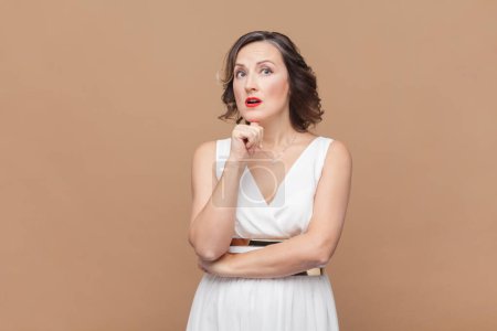 Photo for Portrait of thoughtful woman with wavy hair standing holding chin, looking away, thinks about something astonished, wearing white dress. Indoor studio shot isolated on light brown background. - Royalty Free Image