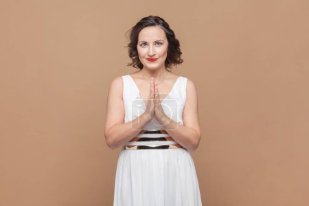 Photo for Portrait of smiling happy middle aged woman with wavy hair standing keeps palms together, looking at camera, wearing white dress. Indoor studio shot isolated on light brown background. - Royalty Free Image