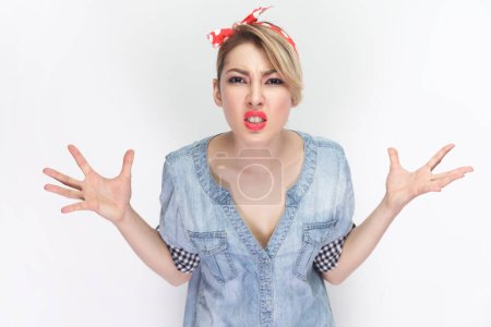 Photo for Angry annoyed woman in blue denim shirt and red headband gestures with annoyance, screams loudly, expresses irritation, reproaches partner in betrayal. Indoor studio shot isolated on gray background. - Royalty Free Image