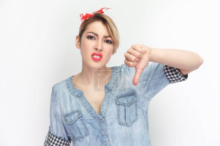 Photo for Portrait of sad upset attractive blonde woman wearing blue denim shirt and red headband standing showing thumb down, dislike gesture. Indoor studio shot isolated on gray background. - Royalty Free Image