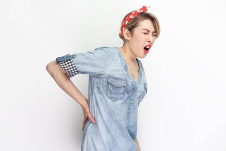 Photo for Portrait of sick blonde attractive woman wearing blue denim shirt and red headband standing touching her painful back, frowning face from terrible pain. Indoor studio shot isolated on gray background. - Royalty Free Image