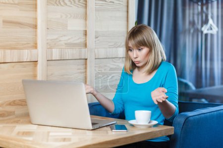 Photo for Portrait of confused uncertain young woman with blonde hair in blue shirt sitting at table and working on laptop, shrugging shoulders, looks doubtful and puzzled. Indoor shot, cafe background. - Royalty Free Image