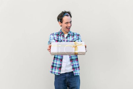 Photo for Positive man with festive mood standing giving present box, celebrating friend's birthday, congratulating, wearing blue checkered shirt and headband. Indoor studio shot isolated on gray background. - Royalty Free Image