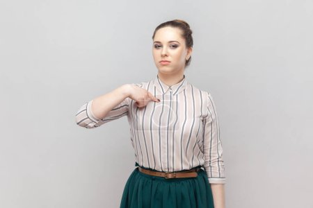Photo for Portrait of strict bossy serious woman wearing striped shirt and green skirt standing pointing at herself with proud expression, looks arrogant. Indoor studio shot isolated on gray background. - Royalty Free Image