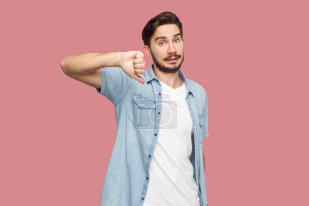 Photo for Portrait of bearded man in blue casual style shirt standing assess project, shows sign of dislike, looks with negative expression and disapproval. Indoor studio shot isolated on pink background. - Royalty Free Image
