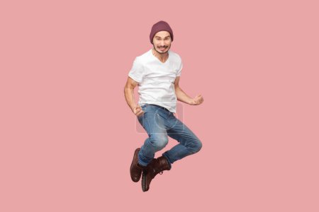 Photo for Portrait of extremely happy joyful bearded man in white T-shirt and beany hat jumping with clenched fists, rejoicing, celebrating victory. Indoor studio shot isolated on pink background. - Royalty Free Image