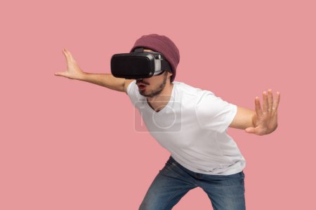 Photo for Portrait of amazed wary bearded man in white T-shirt and beany hat, wearing vr, playing in headset, trying to avoid monster in video game. Indoor studio shot isolated on pink background. - Royalty Free Image