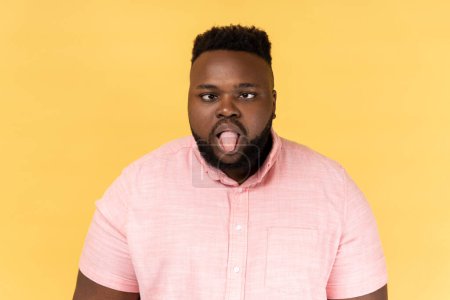 Photo for Portrait of disobedient crazy funny man wearing pink shirt showing tongue out and crossed his eyes displeased, behaving unruly naughty. Indoor studio shot isolated on yellow background. - Royalty Free Image