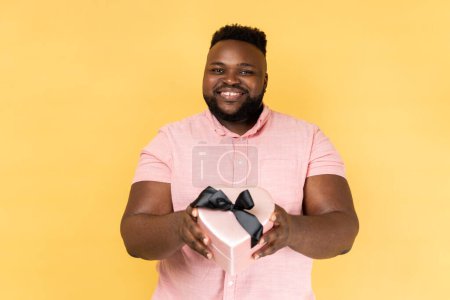 Photo for Portrait of joyful man wearing pink shirt giving heart shaped gift box to camera with excited smile, greeting on holiday and sharing present. Indoor studio shot isolated on yellow background. - Royalty Free Image